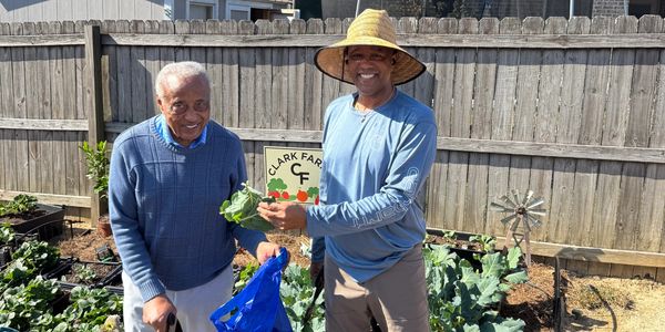 Two men smiling in a garden with fresh vegetables at Clark Farm.