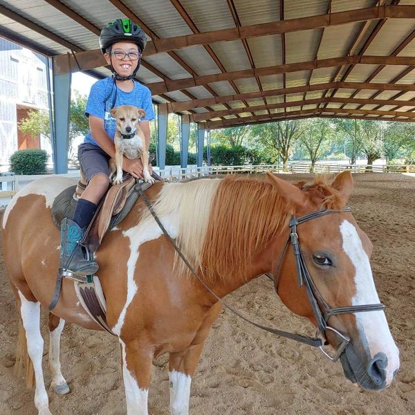 A boy with MSS on horseback. He is smiling and a small dog is sitting with him in the saddle.