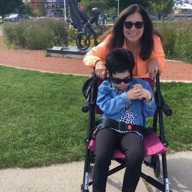 A woman with MSS sits in a wheelchair. Her mother stands behind her. Both are wearing sunglasses.