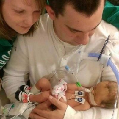 A newborn with MSS is being held by her father in the NICU as her mother looks over his shoulder.