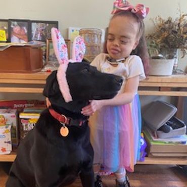 A girl with MSS stands next to her service dog. The dog is black and wearing pink bunny ears.
