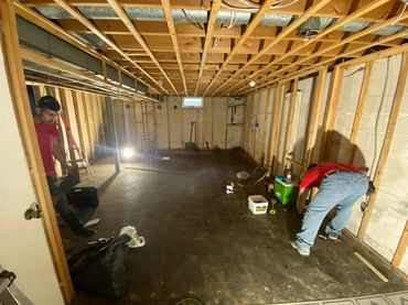 Two men work on renovating an unfinished basement with exposed wooden beams and concrete walls.