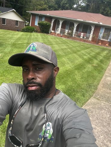 Man in gray shirt and cap taking a selfie in front of a well-manicured lawn.