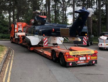 A helicopter is being transported on a large orange flatbed truck through a forested area.