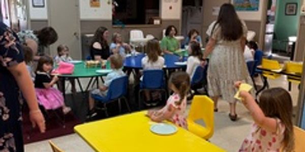 Children sitting and standing around tables in a classroom setting with adults present.