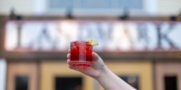 A hand holding a red cocktail with a lime wedge in front of a Landmark sign.