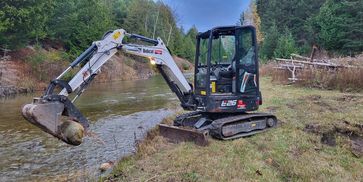 A Bobcat excavator lifting a large rock by a river.