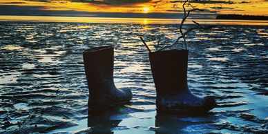 A pair of black rubber boots stand alone on a tidal flat at sunset with driftwood sticking out of th