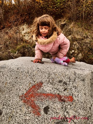 A girl wearing her pink hooded coat backwards kneels on a large boulder and looks down at a red arro