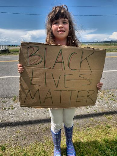 a girl in blue rubber boots stands by a roadside and holds a carboard sign that reads "Black Lives M