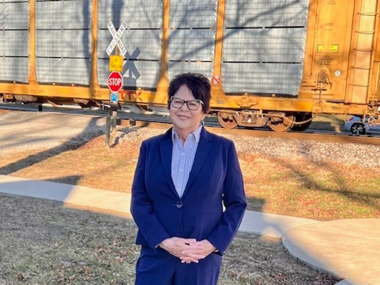 Woman in blue suit standing on sidewalk near railroad tracks with a train passing by.