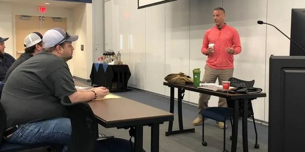 A man in a red shirt speaks to seated attendees in a classroom or seminar setting.