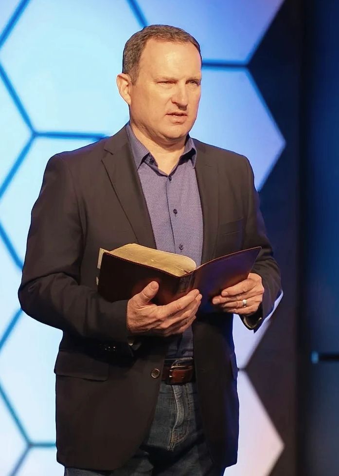 Man in blazer reading from a book on stage with hexagonal background.