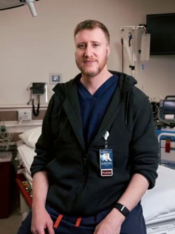 A male physician in a hospital room, wearing a badge and casual medical attire.