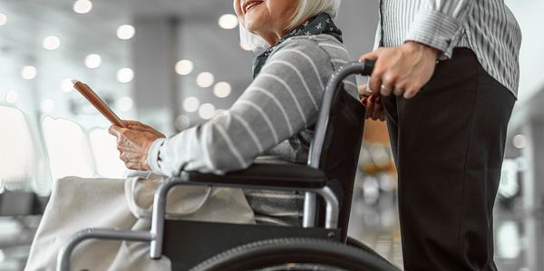 An elderly woman in a wheelchair smiling while holding a tablet, assisted by a person standing behind her.