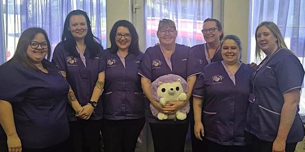 Seven women in matching uniforms smiling indoors, one holding a stuffed hedgehog.