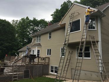 Two workers on ladders repairing the exterior of a large beige house.