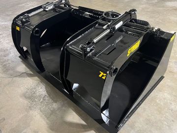 Black heavy-duty grapple bucket attachment with hydraulic cylinders on a concrete floor.