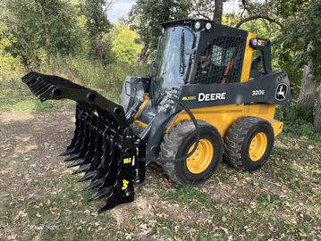 John Deere 320G skid steer loader with a heavy-duty grapple attachment in a wooded area.
