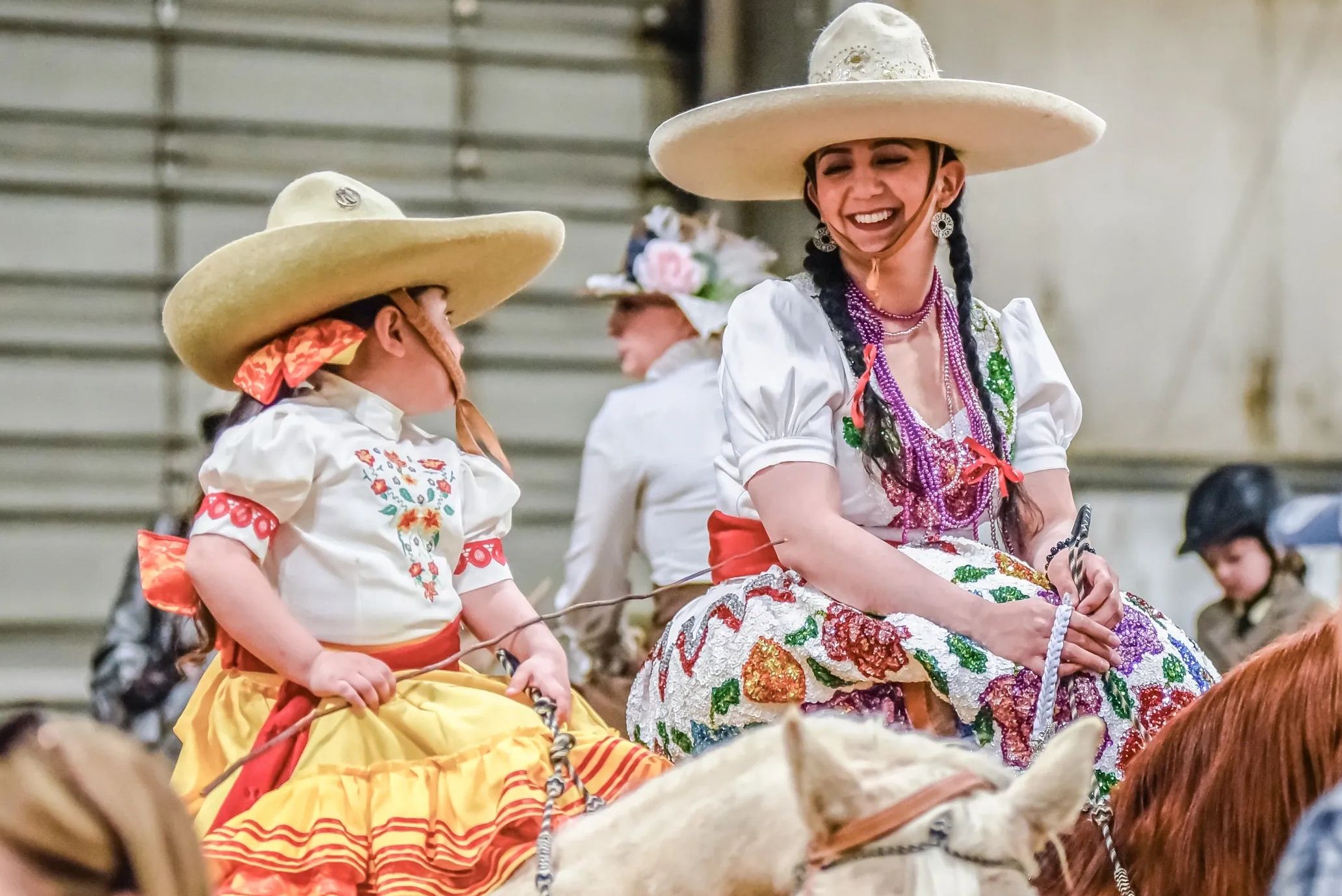 Two women in traditional Mexican attire smiling while riding horses.