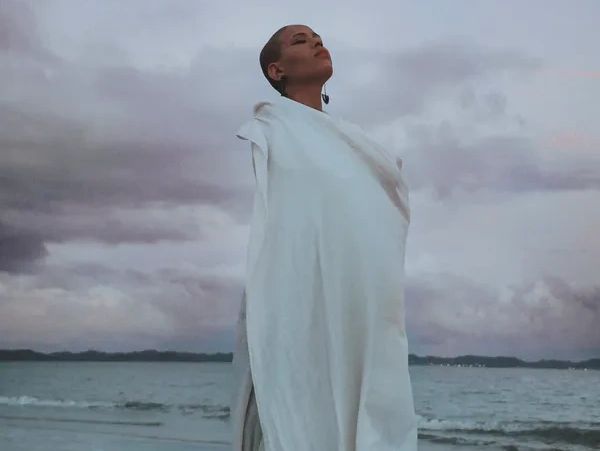 Person in white robe standing by the sea with eyes closed.
