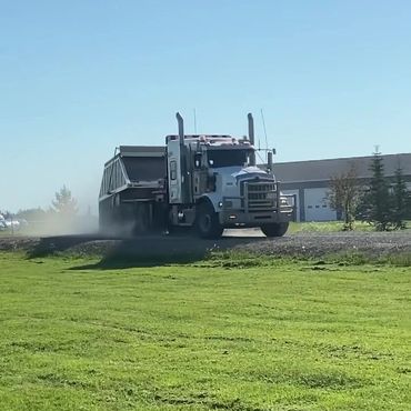 White semi-truck with trailer driving on a gravel path near a green field.