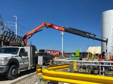 Workers in safety gear operate a crane truck near industrial tanks on a clear day.