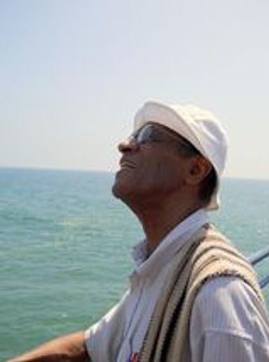 Elderly man gazing thoughtfully at the ocean while wearing a white hat and sunglasses.