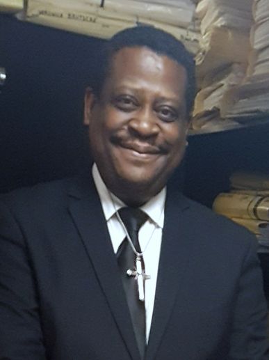 Man in a black suit with a cross necklace smiling.