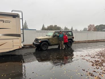 Two people stand next to a Jeep towing a camper on a rainy day.