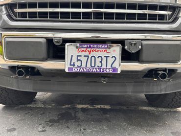 Close-up of a California license plate on a Ford vehicle with 'Light The Beam' tag.