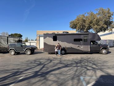 Couple with a dog standing next to a large RV and a Jeep in a parking lot.