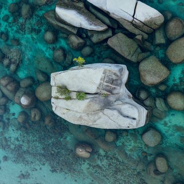 Large white rock with small trees surrounded by clear turquoise water and smaller rocks.