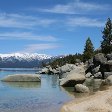 Clear lake with rocks and snowy mountains in the background under a blue sky.