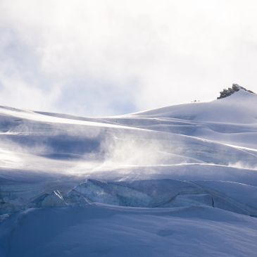 Snow-covered mountain slopes with wind blowing snow into the air.