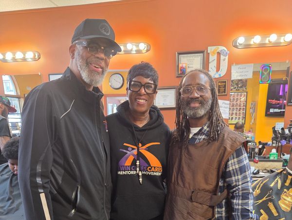 Three smiling adults posing inside a barbershop with warm lighting.