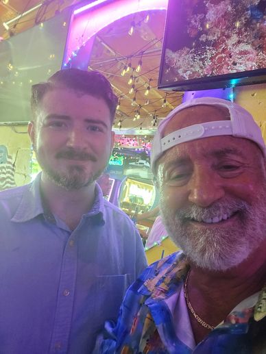 Two men smiling together in a colorful, festive indoor setting.