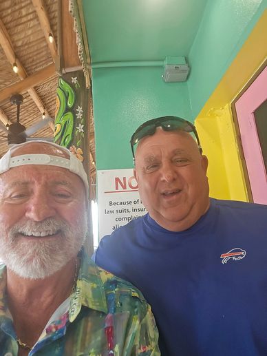 Two smiling men posing indoors with colorful walls and a ceiling fan.