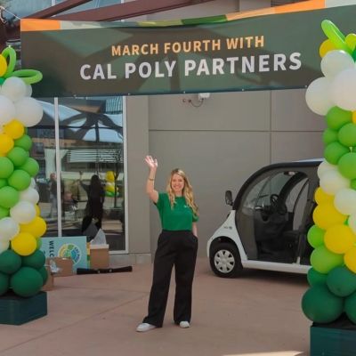 A woman waves under Cal Poly Partners banner with balloon decorations.