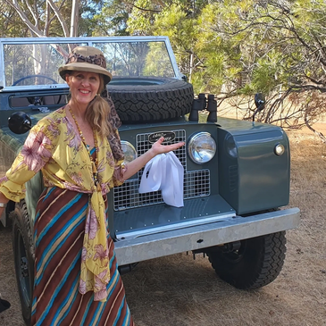 Woman smiling beside a vintage Land Rover in a sunny, wooded area.