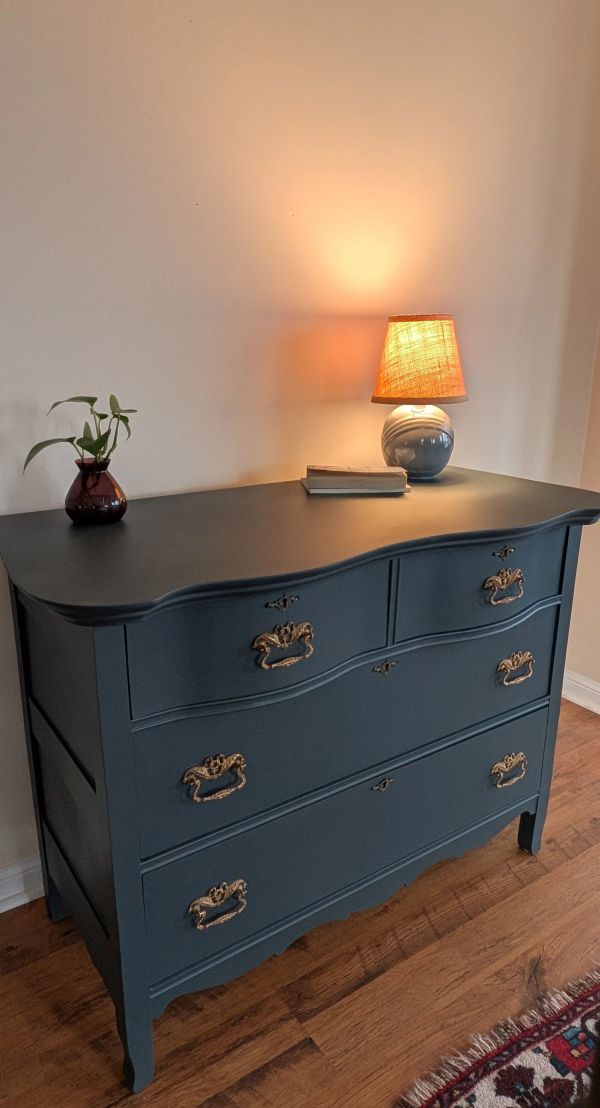 Blue vintage dresser with ornate handles, lamp, and small plant on top.