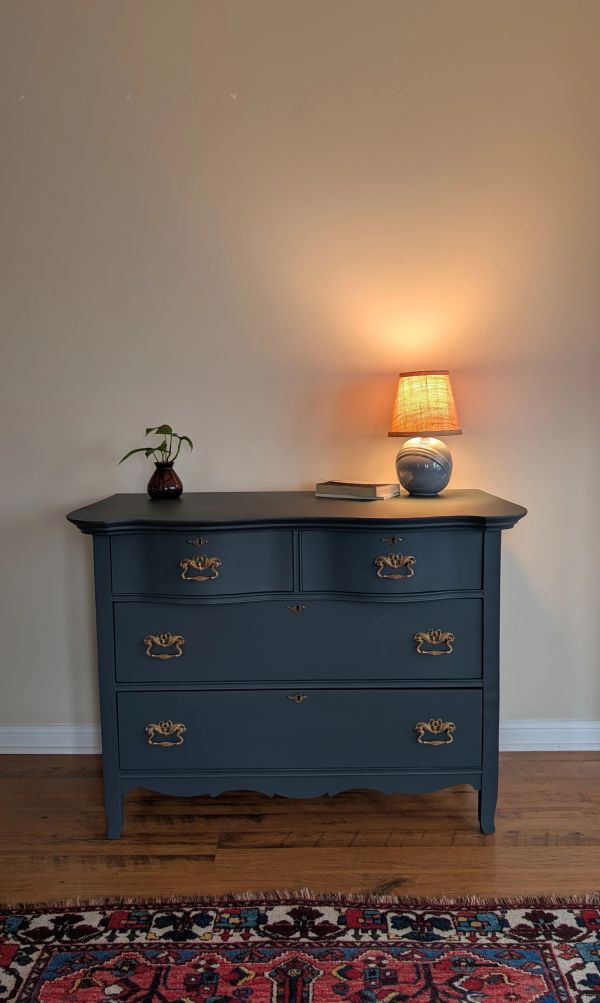 Blue vintage dresser with brass handles, topped with a small lamp and plant.