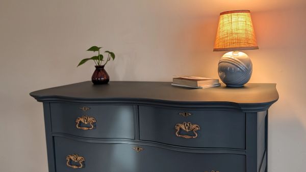A blue dresser with vintage brass handles, decorated with a small plant and a lit lamp.