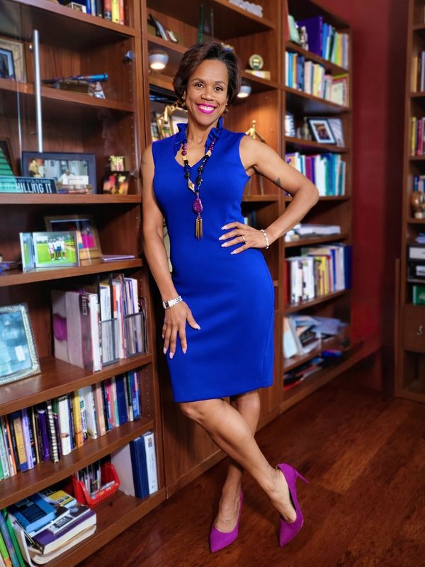 Woman in a blue dress and purple heels poses in a library or study.