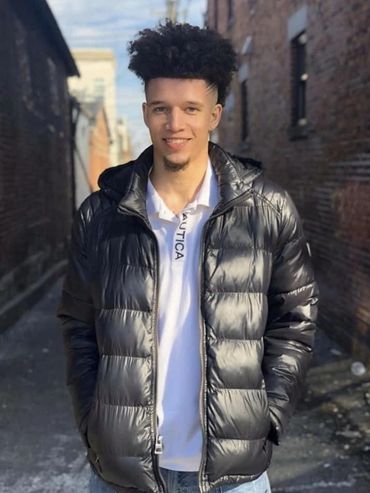 Chandler Bussey poses in an alley wearing a black bubble jacket and a white Nautica polo shirt.