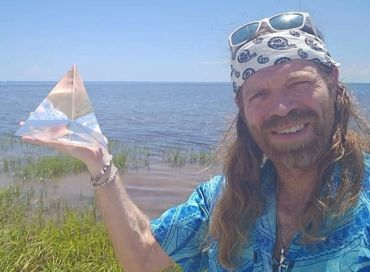 Man wearing a bandana holds a small pyramid-shaped object by a lakeside.