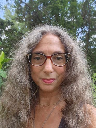 Woman with long gray hair and glasses outdoors in a natural setting.