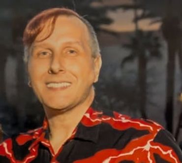A smiling man with light hair in a red and black patterned shirt.