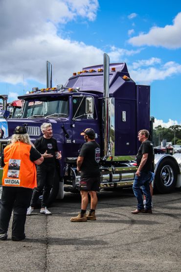 People talking beside a large purple semi-truck under a partly cloudy sky.