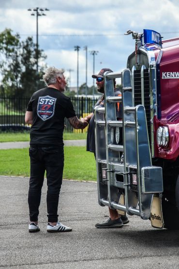 Two men shaking hands beside a large red Kenworth truck outdoors.